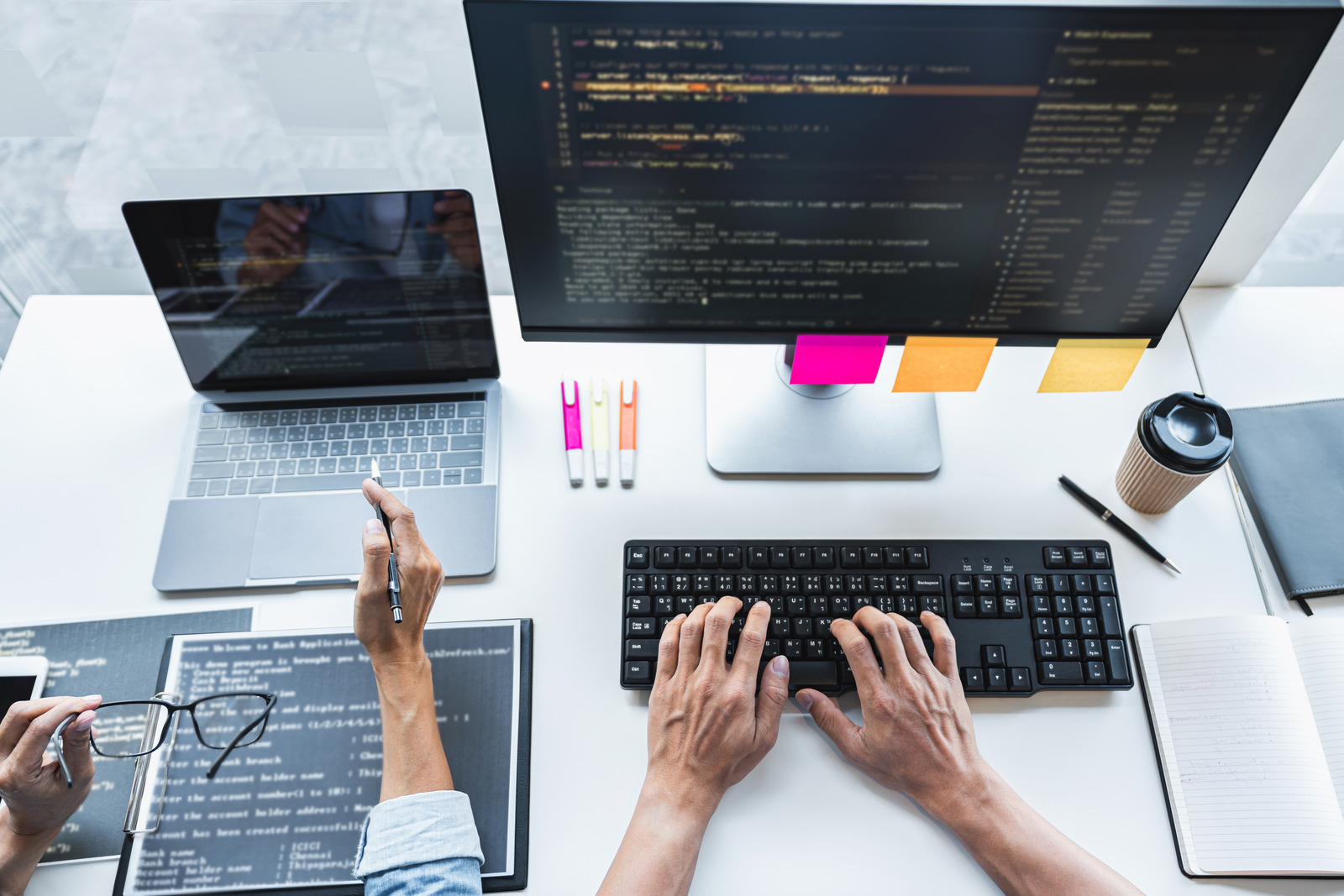 Duas pessoas programando um site. Uma delas em um notebook e outra em um computador de mesa. Ambas estão com diversos códigos na tela.